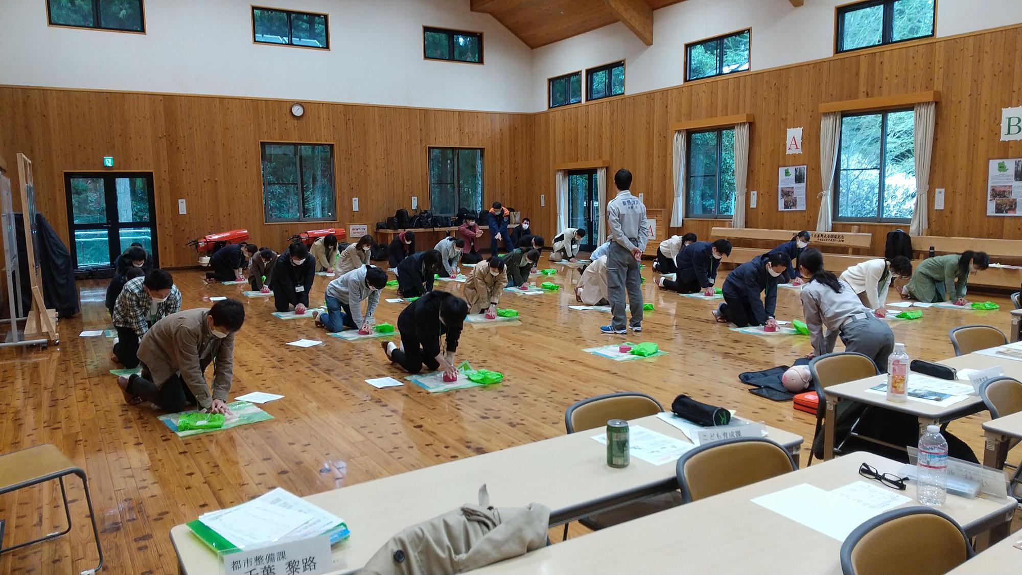 フローリングの広い室内で、大勢の参加者が床に膝をつき、心肺蘇生法などの応急処置訓練を受けている様子の写真