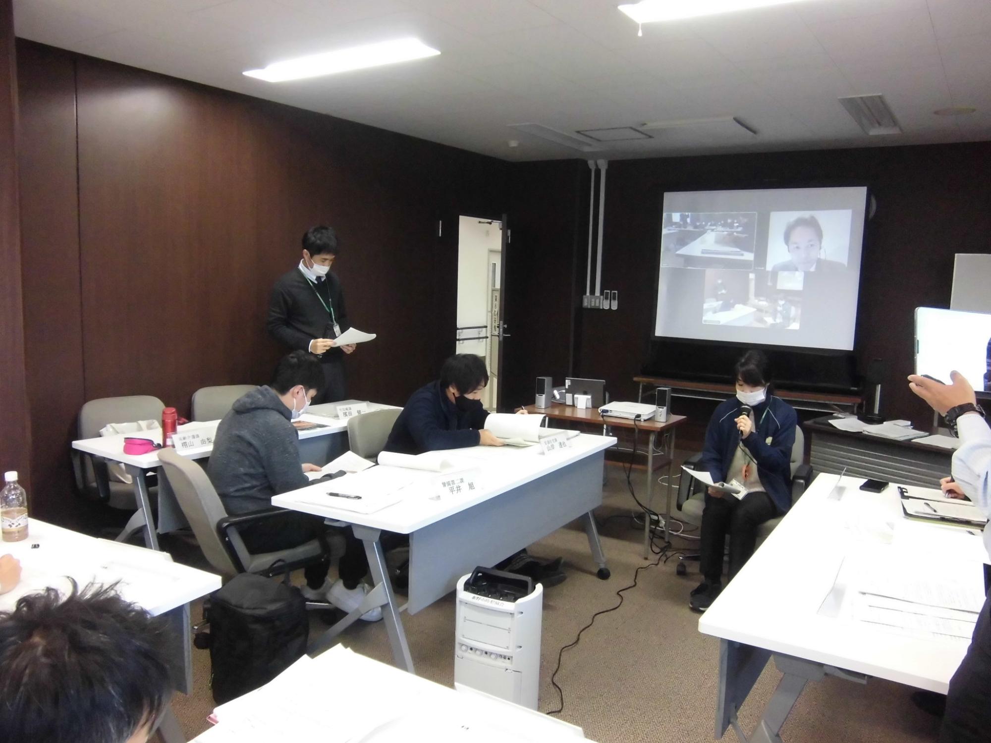 数人がテーブルに向かって資料を読み込み、テーブルの間の椅子に座った女性がマイクを持って発言し、背後のスクリーンにはオンライン参加者の顔が映し出されている研修の様子の写真