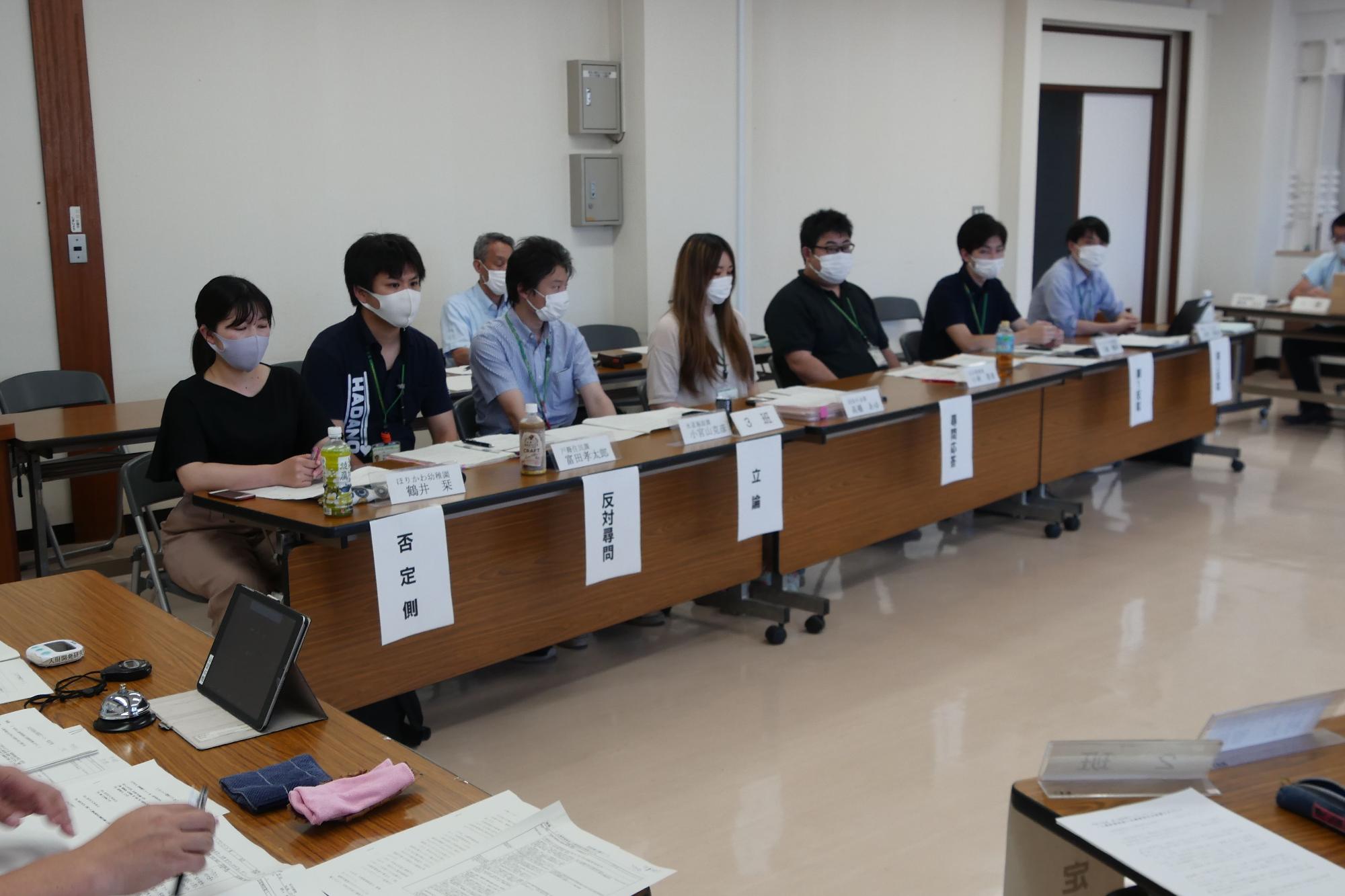 会議用テーブルに並んで座った参加者、それぞれの前に「否定側」「反対尋問」「立論」といった役割分担を示す札が置かれているディベートの様子の写真