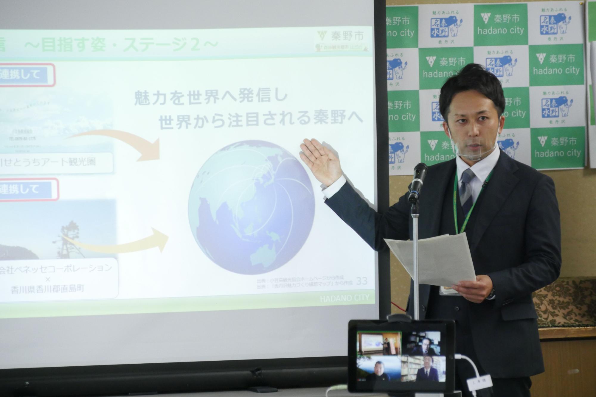 スーツ姿の男性が、資料が映し出されたスクリーンを指しながら、資料を片手に説明をしている様子の写真