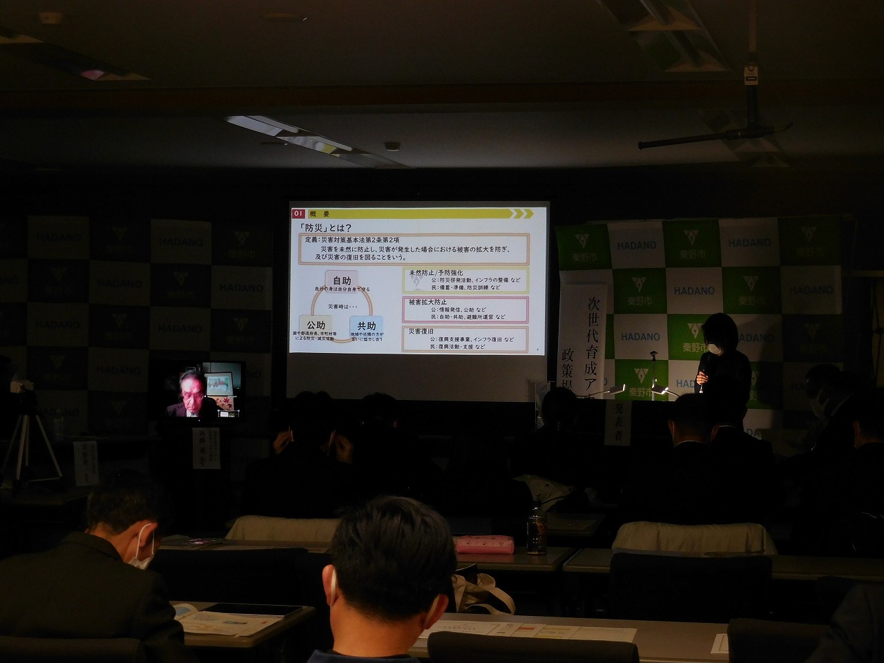 照明を落とした会場で、スクリーンに防災対策に関する資料が映し出され、着席した出席者たちが注目している様子の写真