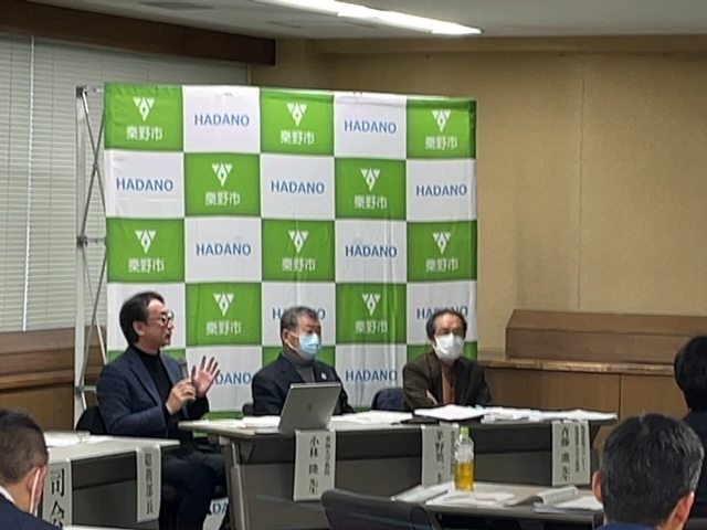 秦野市のバックパネルの前で、3人の男性がテーブルに着席しており、左端の男性が手振りをつけて発言している会議の様子の写真