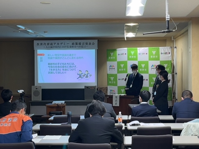 「次世代育成アカデミー 政策提言発表会」の会場で、スーツ姿の発表者たちが聴衆に向かって提言を行っている様子の写真