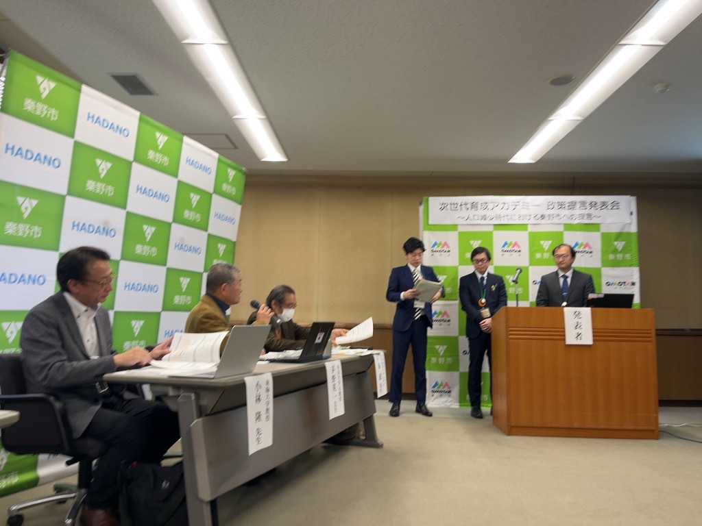 「次世代育成アカデミー 防災提案発表会」の看板がある会場で、市長や市職員らが緑と白のチェック柄の背景幕の前に立つなか、数名の発表者と審査員らしき人々が壇上とテーブルについている会議の様子の写真