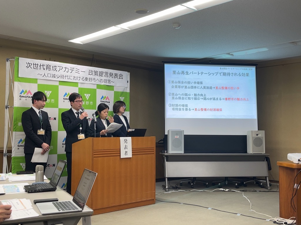 「次世代育成アカデミー 政策提言発表会」の舞台で、スーツ姿の4人の発表者が檀上に立ち、スクリーンには「里山再生パートナーシップで期待される効果」として3つの効果が箇条書きで示されている発表会の写真