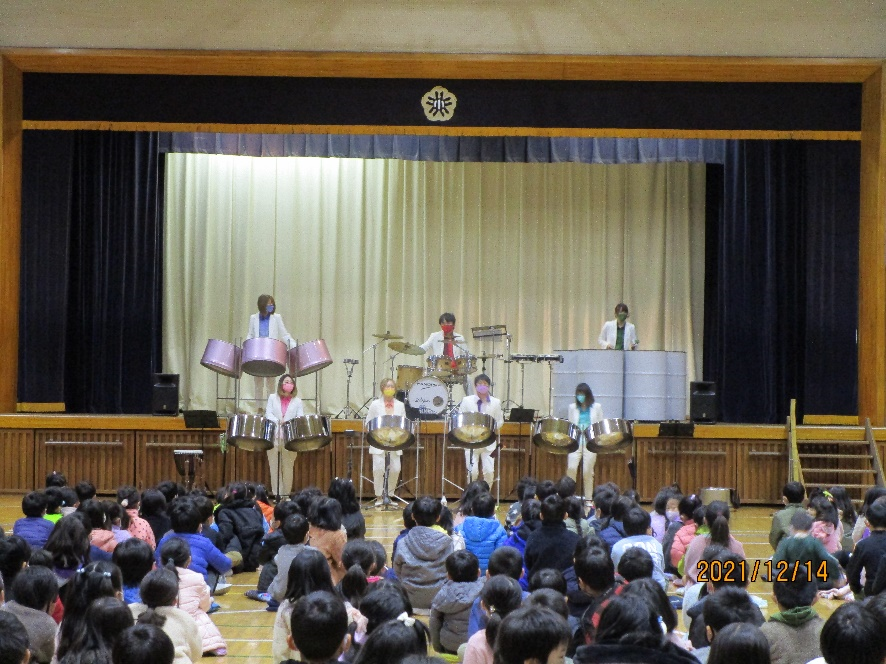 学校の体育館の舞台で白い衣装とカラフルなマスクをつけた7人の演奏者が、スティールパンを使い演奏しているのを床に座り鑑賞している児童の写真