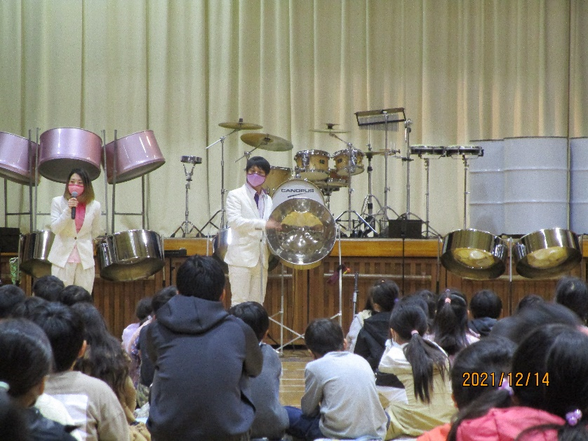 体育館の舞台の下で白いスーツとマスクを着用した女性がマイクを持って話をし、男性が特徴的な形状のスティールパンを床に座っている児童に見せている写真