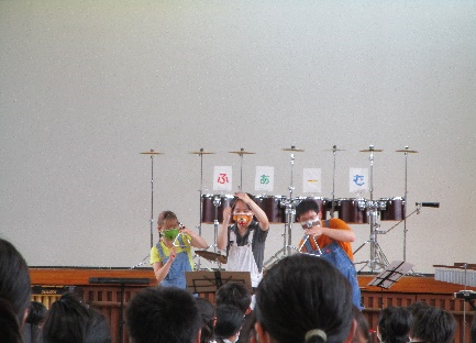 体育館の舞台の下でオーバーオール姿の3人の方がトライアングルを持ち叩いているのを児童が鑑賞している写真