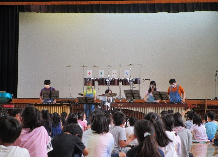 体育館の舞台の下で5人の方が楽器を演奏しているのを床に座って鑑賞している児童を後ろから写した写真