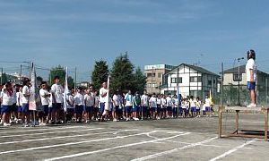 晴天の下、校庭のトラック上に整列した大勢の中学生が、壇上に立つ生徒の開会の言葉を聞いている体育祭開会式の写真