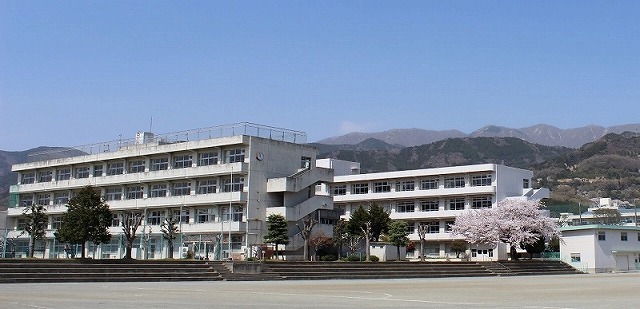 遠くの山々を背景に、グラウンド奥に立つ、白い四階建て校舎が2棟並んだ校舎の外観写真