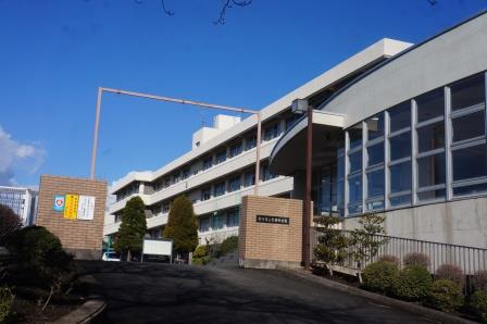 円弧状のデザインで大きな窓が特徴的な建物の奥には白色の四階建ての校舎を門柱も写るように全体的に写した大根中学校の外観写真